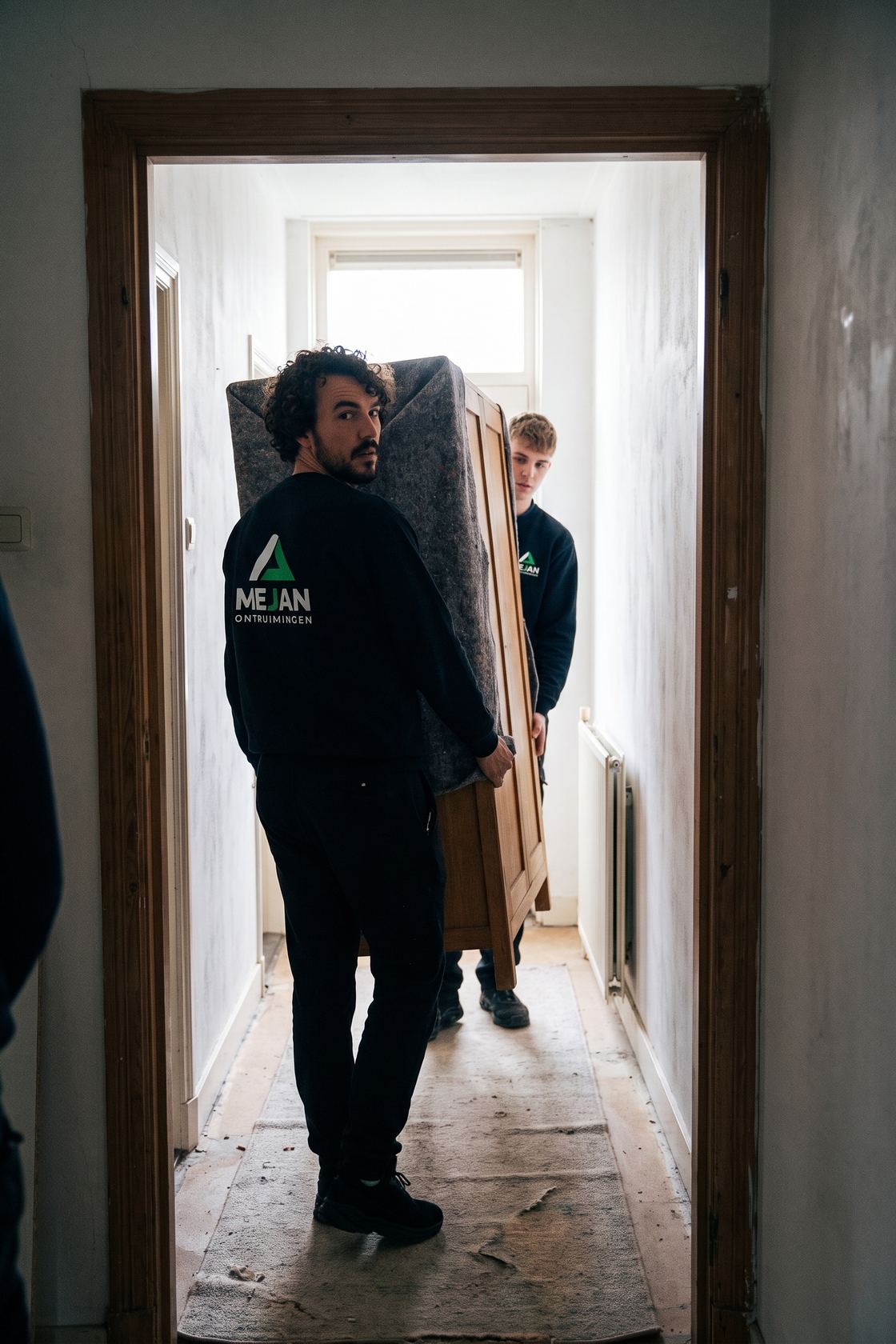 Twee medewerkers dragen een kast in dekens door de gang, gezien door de deurpost