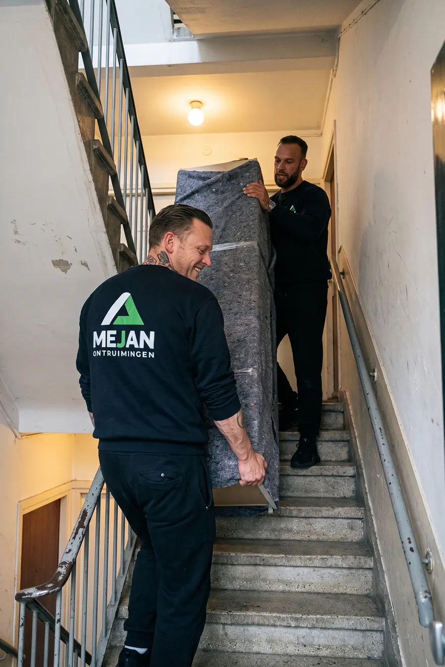 Twee medewerkers dragen een in dekens gepakte kast door het trappenhuis